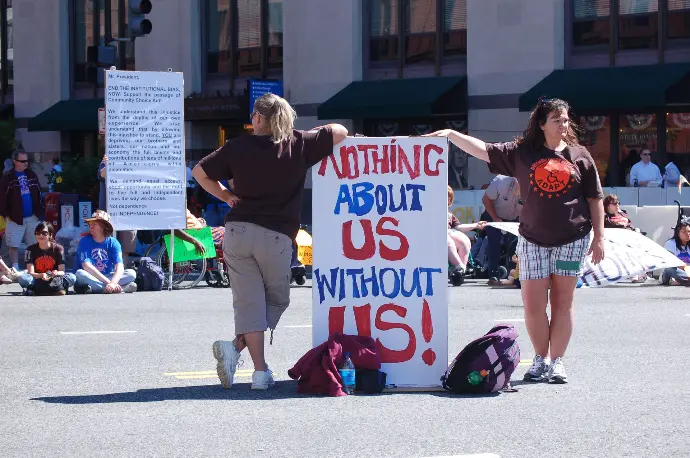 Image of two demonstrators in the middle of a road holding a sign that says "nothing about us without us!"