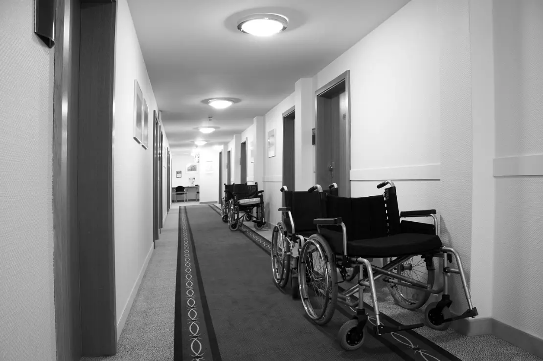 A stock photo is used adjacent to the NHTD article. It displays a long, narrow indoor hallway in a healthcare or assisted living facility, shown in black and white. The corridor has smooth, light-colored walls with several closed doors on both sides, each with a handle and small signage. Overhead, evenly spaced round ceiling lights illuminate the space.  Along the right side of the hallway, multiple empty wheelchairs are lined up facing slightly toward the center, with metal frames, large rear wheels, and footrests visible. The floor is covered with a carpet that includes a darker central strip with a subtle patterned design running down the length of the corridor.  At the far end of the hallway, a small desk or table and a chair are visible against the back wall, adding depth and perspective. The overall scene feels quiet, orderly, and institutional.
