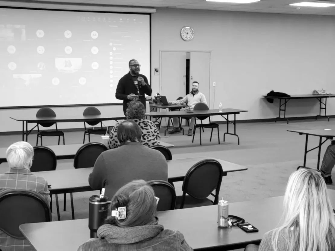 Above this article, a photo is shown of the Reverend Doctor Damond A. Wilson, a Black man in his thirties with a beard and glasses wearing dark clothing, addressing STIC.  He is standing in front of a projection screen showing a computer desktop and several tables and chairs.  To Reverend Wilson’s right, a White man in a light top with a heavy beard operates a desktop beneath a clock reading 9:32.  In the foreground we see the backs of the heads of audience members.